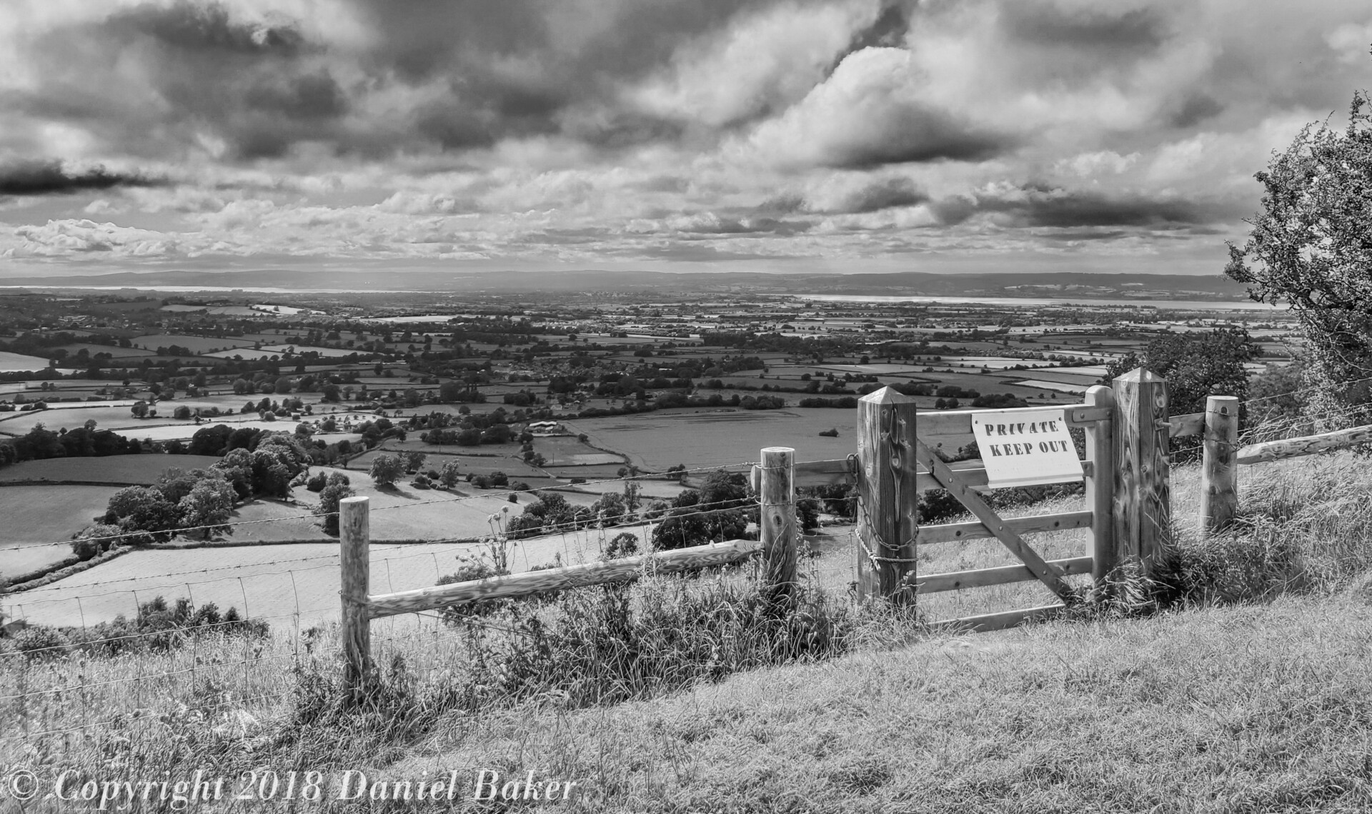 A black and white fence photograph of a fence with keep out written on it, behind which the land drops suddenly with a rural landscape in view reaching off into the distance