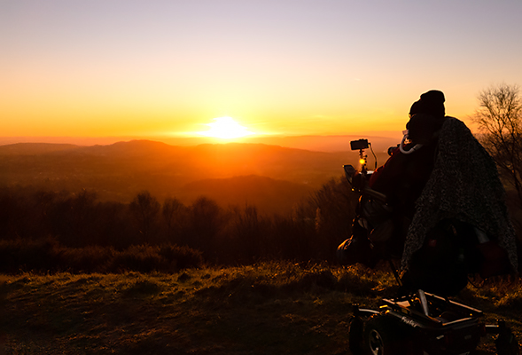 Daniel Baker sitting in his wheelchair taking a photograph of a sunset over the Malvern hills