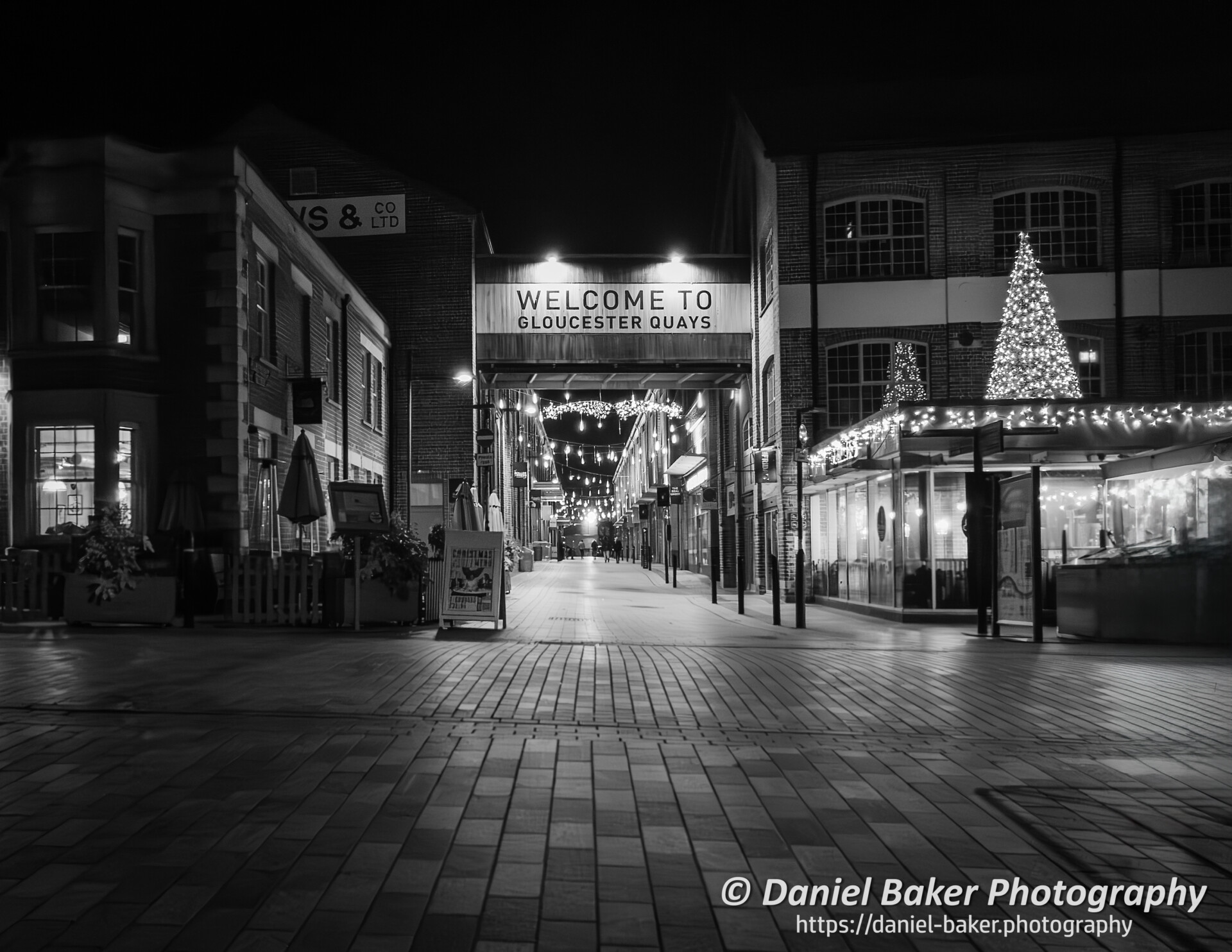 A black and white photo taken at night taken at Gloucester Quays showing shops lit by Christmas Decorations