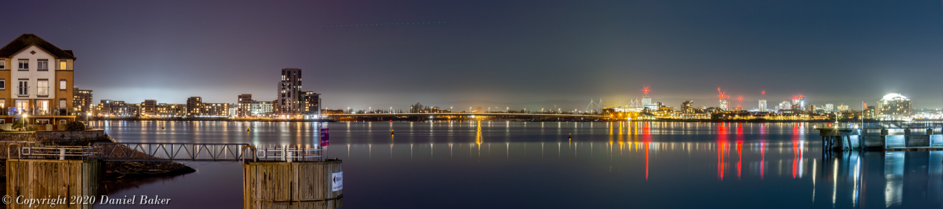 A nighttime view of Cardiff bay, with the vast city of coloured lights on the far side being reflected in the water