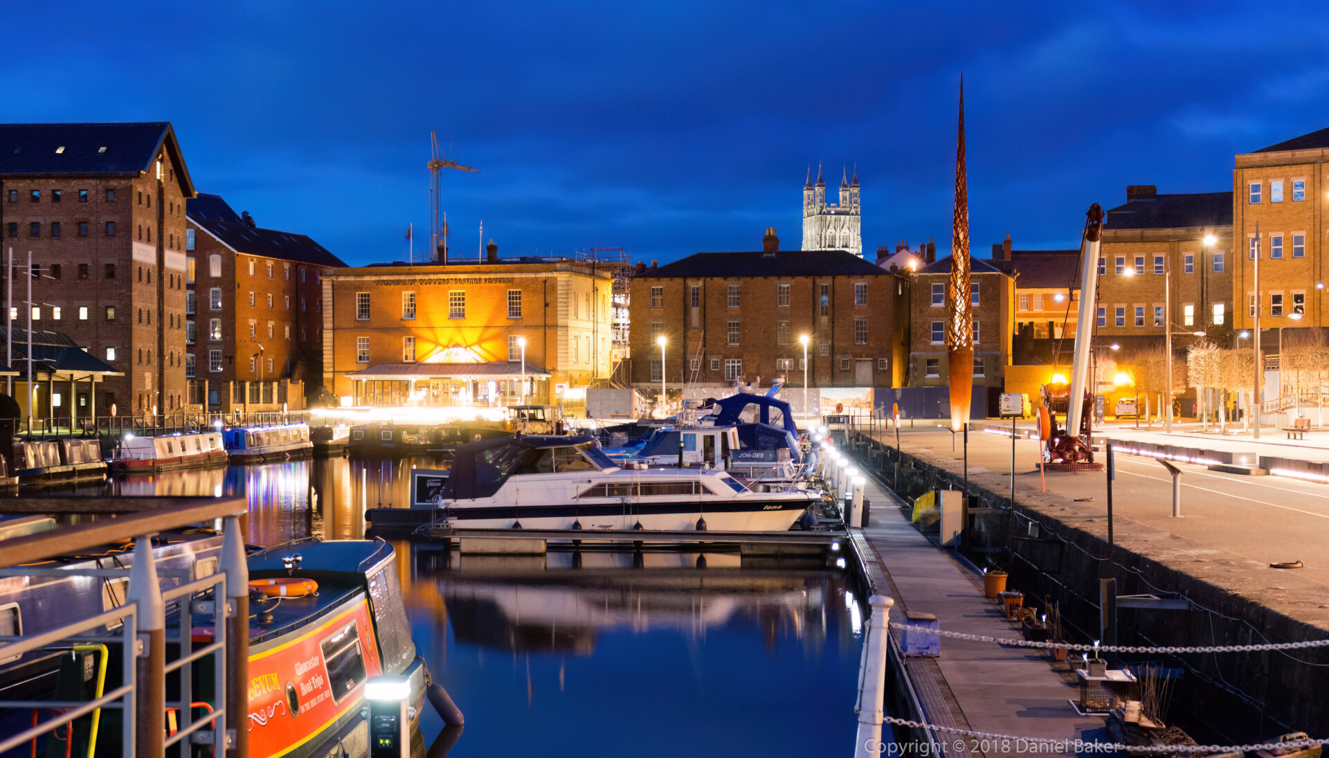 Nighttime view of Gloucester docks, old dockyard buildings bordering the water and the tower of Gloucester Cathedral visible above rooftops in the distance 