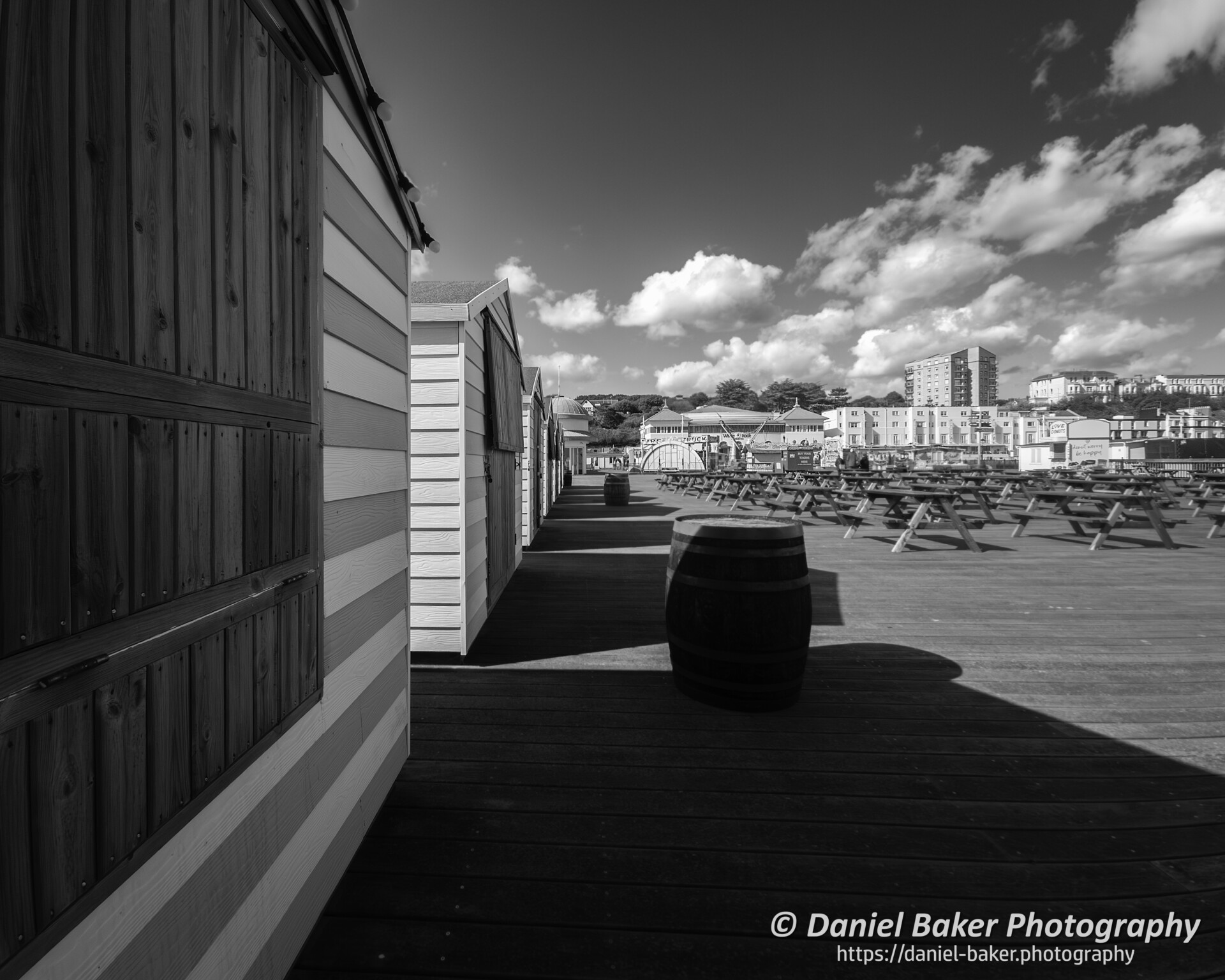 Black and white photo looking down the deck of a piear towards Hastings seafront. Strong shadows being cast by beach huts.