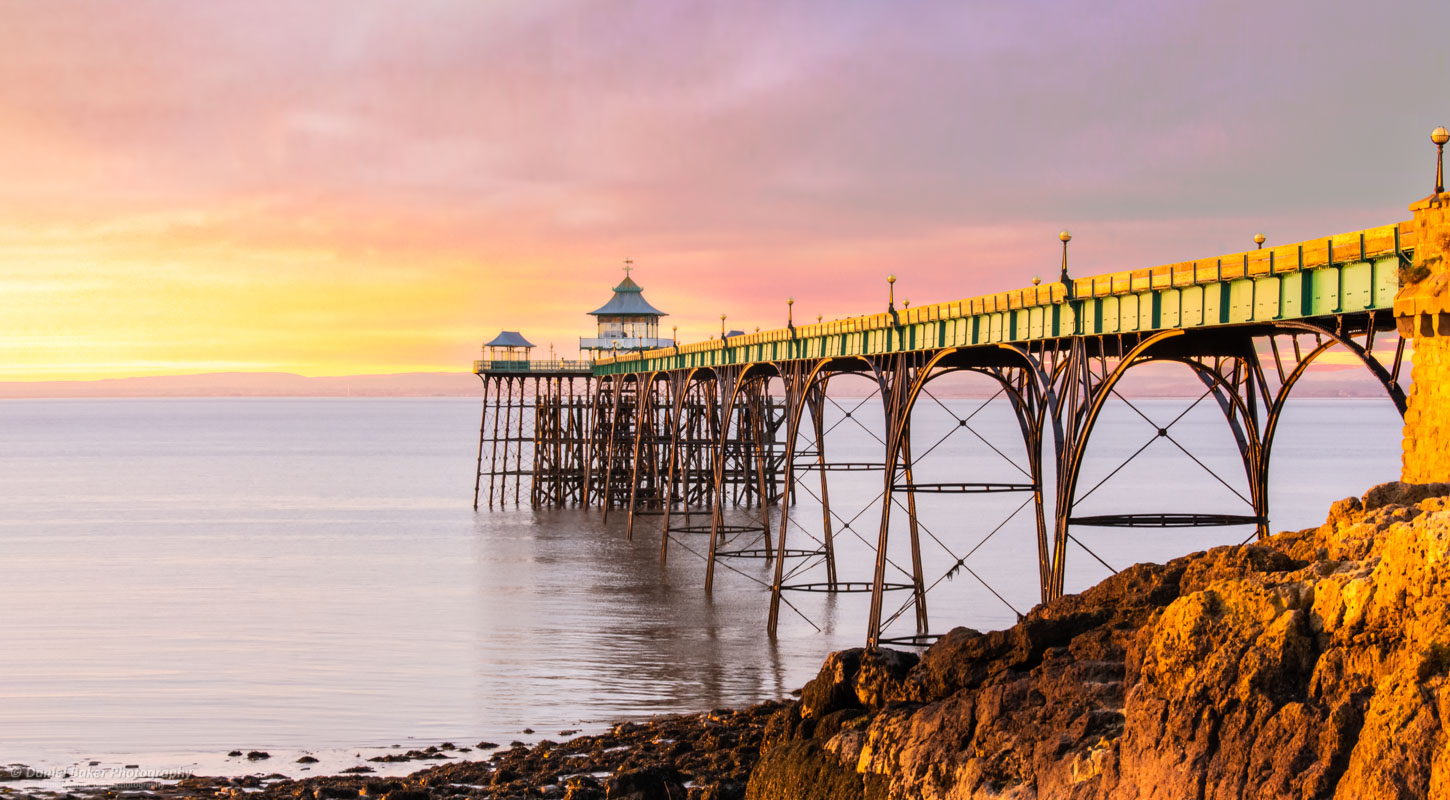 A photograph of a long pier extending into the sea, supported by a series of arches. At the pier’s end, there’s a structure—possibly a pavilion or shelter. The warm hues of the sky suggest a sunset, reflecting in the calm sea.