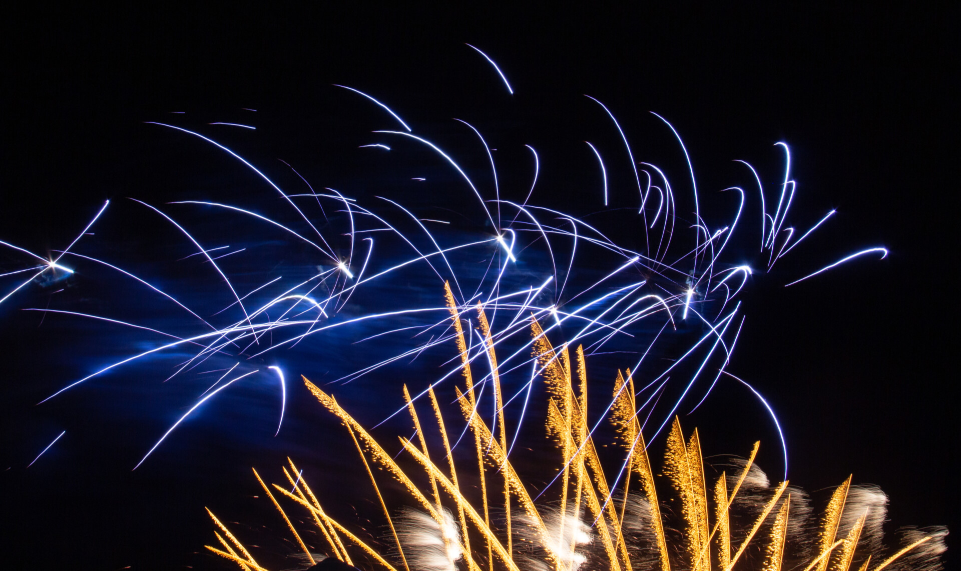 A captivating fireworks display against a night sky. The streaks of light, predominantly blue and gold, burst outward, creating an intricate pattern. The long exposure captures the trails of individual sparks, conveying the excitement and beauty of a fireworks show.