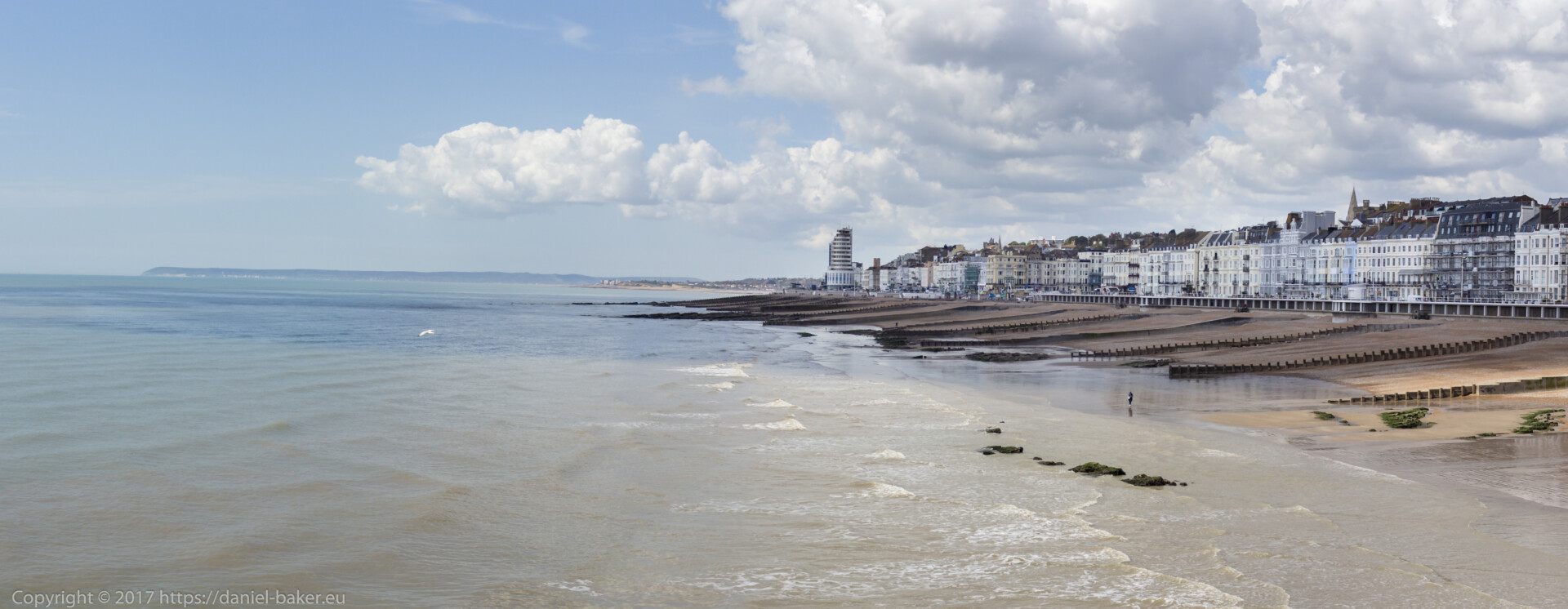 The photograph depicts a panoramic coastal area with a beachfront. Sandy sections alternate with groynes. Along the coastline, there’s a row of buildings, including a distinctive white tower. The calm sea gently laps the shore under a clear sky with scattered clouds. This scene could inspire discussions on coastal architecture, erosion prevention, or simply showcase the area’s natural beauty.