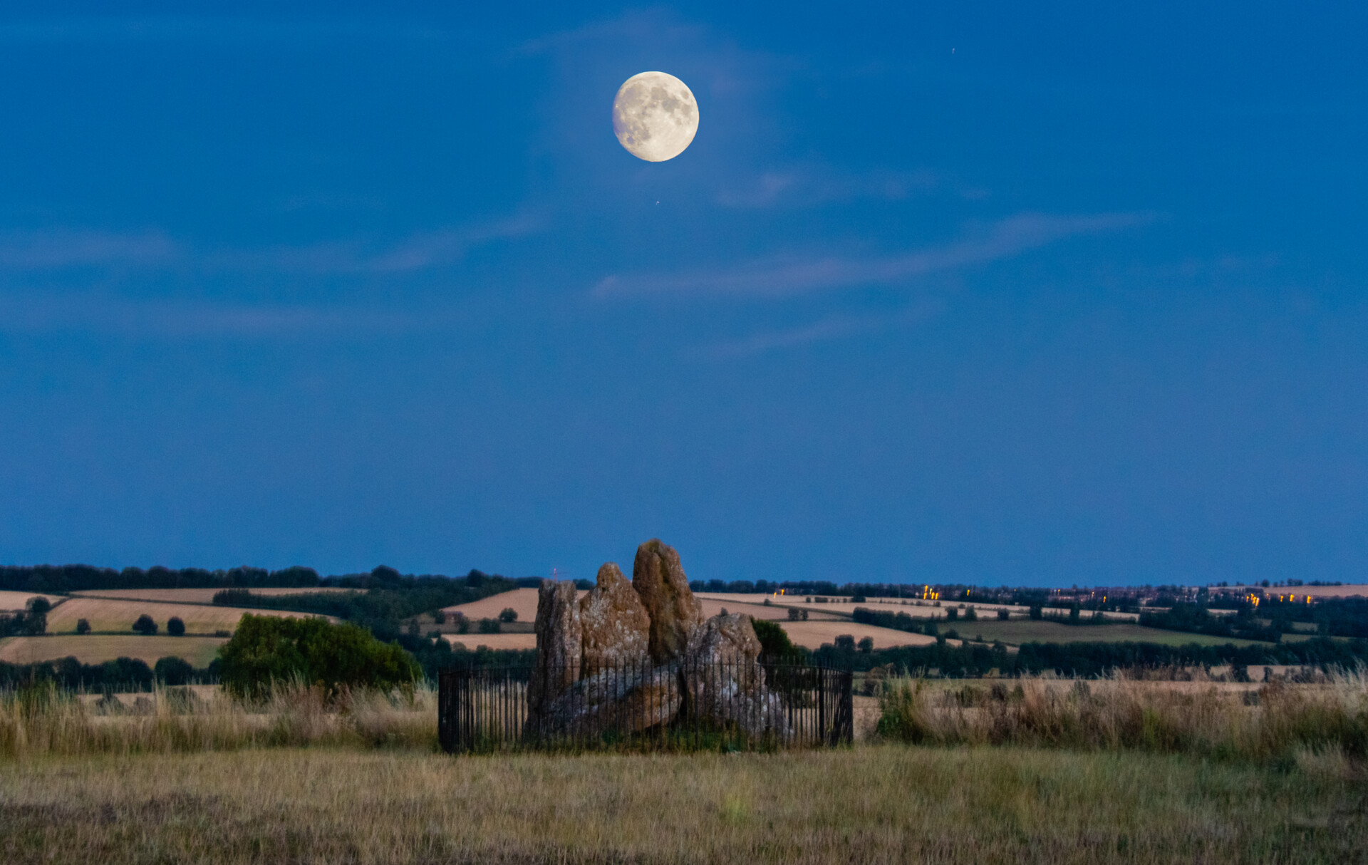 A large, full moon hanging in the sky above a rural landscape during twilight. In the foreground, the striking, jagged rock formation of the whispering Knights neolithic stones are surrounded by a fence. The background features rolling hills with patches of trees and fields, suggesting an agricultural setting. The contrast between the early night sky and the remaining light on the horizon makes this scene captivating, emphasizing the rock formation and creating a serene atmosphere.